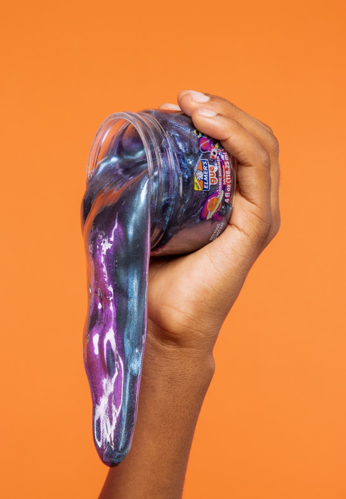 A hand squeezing Cosmic Shimmer slime out of the jar on an orange backdrop