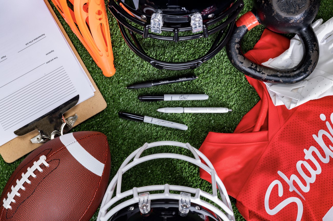 Overhead flat lay of Sharpie pens, helmets, cleats, jersey and a clipboard on field turf